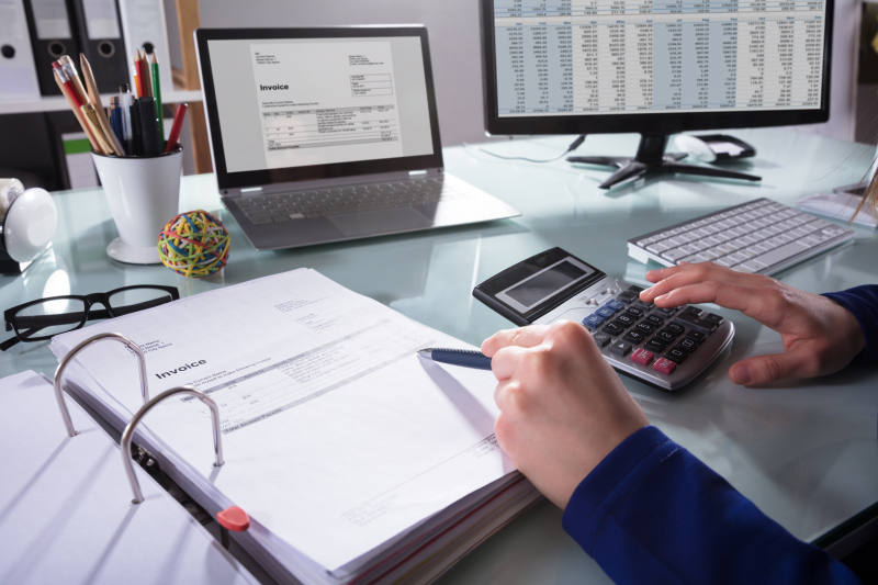 A man is working on a laptop and a PC placed on a white table. He cross-checks the bills and the invoices.